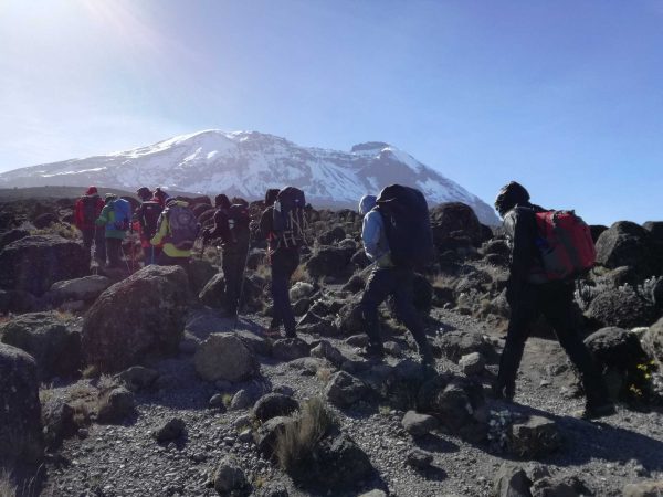 A group of hikers, including porters and guides, making their way up the winding mountain trail on 9 Days Conquer Kilimanjaro Hike via the Northern Circuit Route, with the snow-capped peak looming in the background."