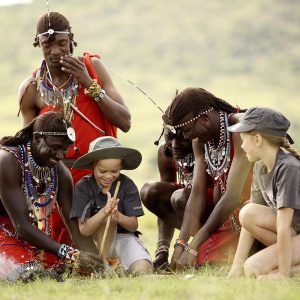 A group of Maasai men gathered around making traditional fire, adorned in colorful clothing and jewelry, preparing a traditional meal together