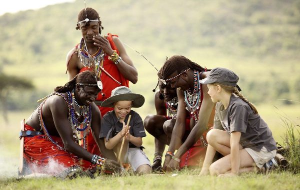 A group of Maasai men gathered around making traditional fire, adorned in colorful clothing and jewelry, preparing a traditional meal together